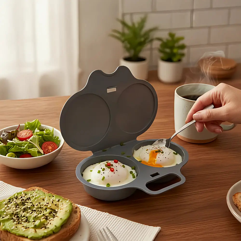 A gray silicone double-egg poacher sits on a wooden table in a bright kitchen setting, with two poached eggs cooking inside. A hand is using a spoon to flip one egg.