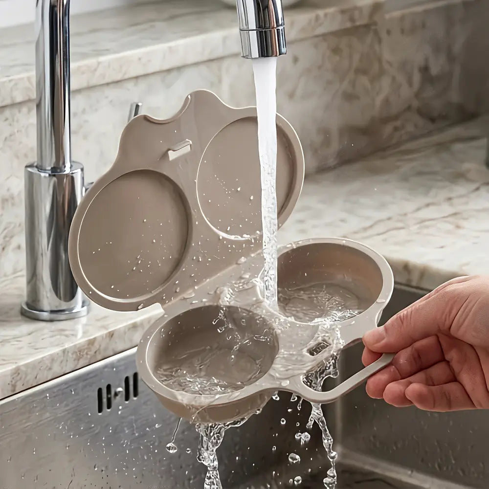 A beige silicone egg cooker with two compartments is shown being rinsed under running water from a kitchen faucet. A person's hand is holding the handle, and a marble kitchen sink and countertop are visible in the background.