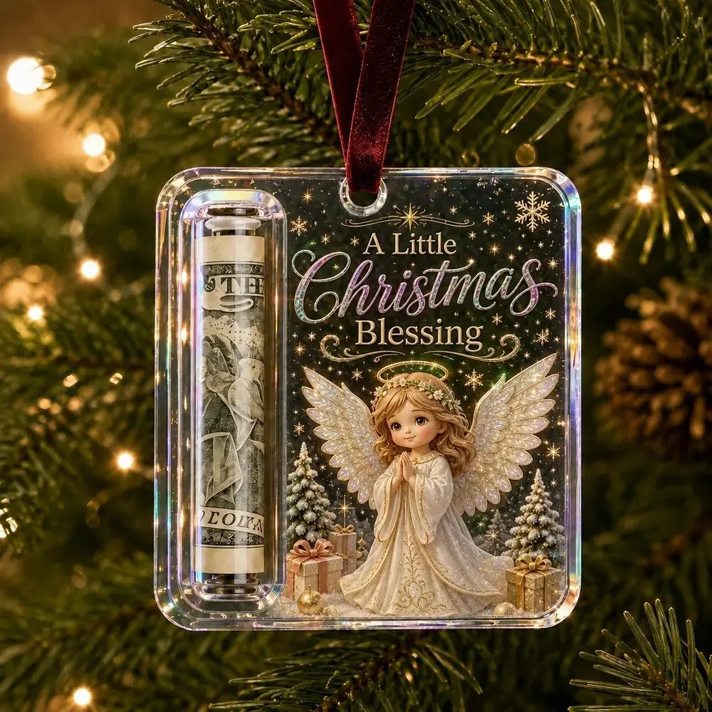 A personalized acrylic Christmas ornament hanging on a Christmas tree. The ornament features a printed illustration of a praying angel, a rolled dollar bill, and the text 'A Little Christmas Blessing' on a dark green background with gold glitter stars.