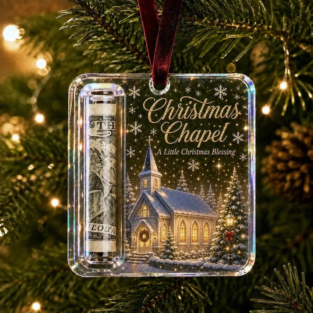 A clear square Christmas ornament hanging on a decorated Christmas tree. The ornament features a printed winter church scene with snow, trees, and the text 'Christmas Chapel' and 'A Little Christmas Blessing,' plus a rolled dollar bill sealed inside a clear tube.