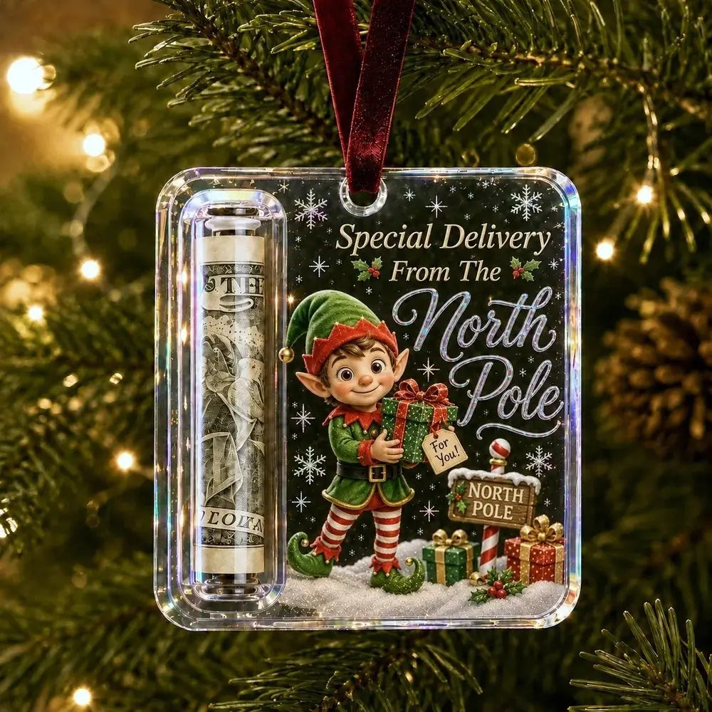 A Christmas ornament hanging on a lit Christmas tree. The ornament is a clear square plastic case with a dark printed holiday design featuring an elf, snow, gift boxes, and the text 'Special Delivery From The North Pole.