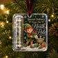 A Christmas ornament hanging on a lit Christmas tree. The ornament is a clear square plastic case with a dark printed holiday design featuring an elf, snow, gift boxes, and the text 'Special Delivery From The North Pole.