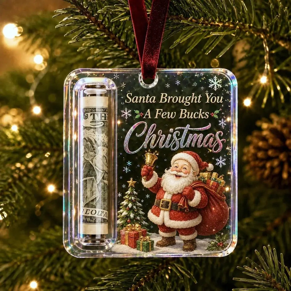 A square Christmas ornament hanging on a decorated tree. The ornament is clear and features a printed design with Santa Claus, a gift sack, a small tree, and the text 'Santa Brought You A Few Bucks Christmas.