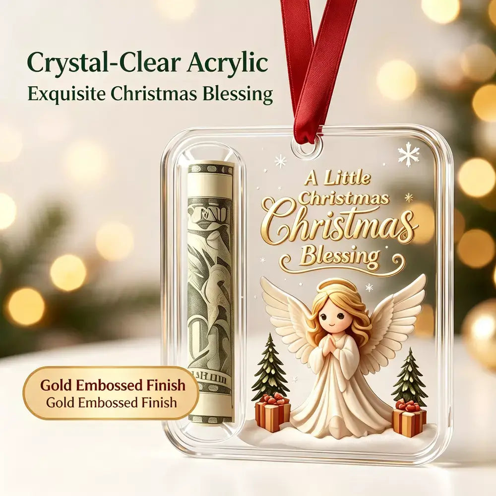 A clear acrylic Christmas ornament with a rolled dollar bill compartment and an embossed angel design with the text 'A Little Christmas Christmas Blessing.' The ornament hangs from a red ribbon against a softly blurred holiday background with warm bokeh lights.