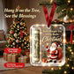 A Christmas tree ornament shown hanging in front of a decorated Christmas tree and wrapped gifts. The ornament is a clear rectangular plastic tag with a red ribbon loop, featuring a rolled dollar bill, Santa Claus, gifts, and the text 'Santa Brought You A Few Bucks Christmas.