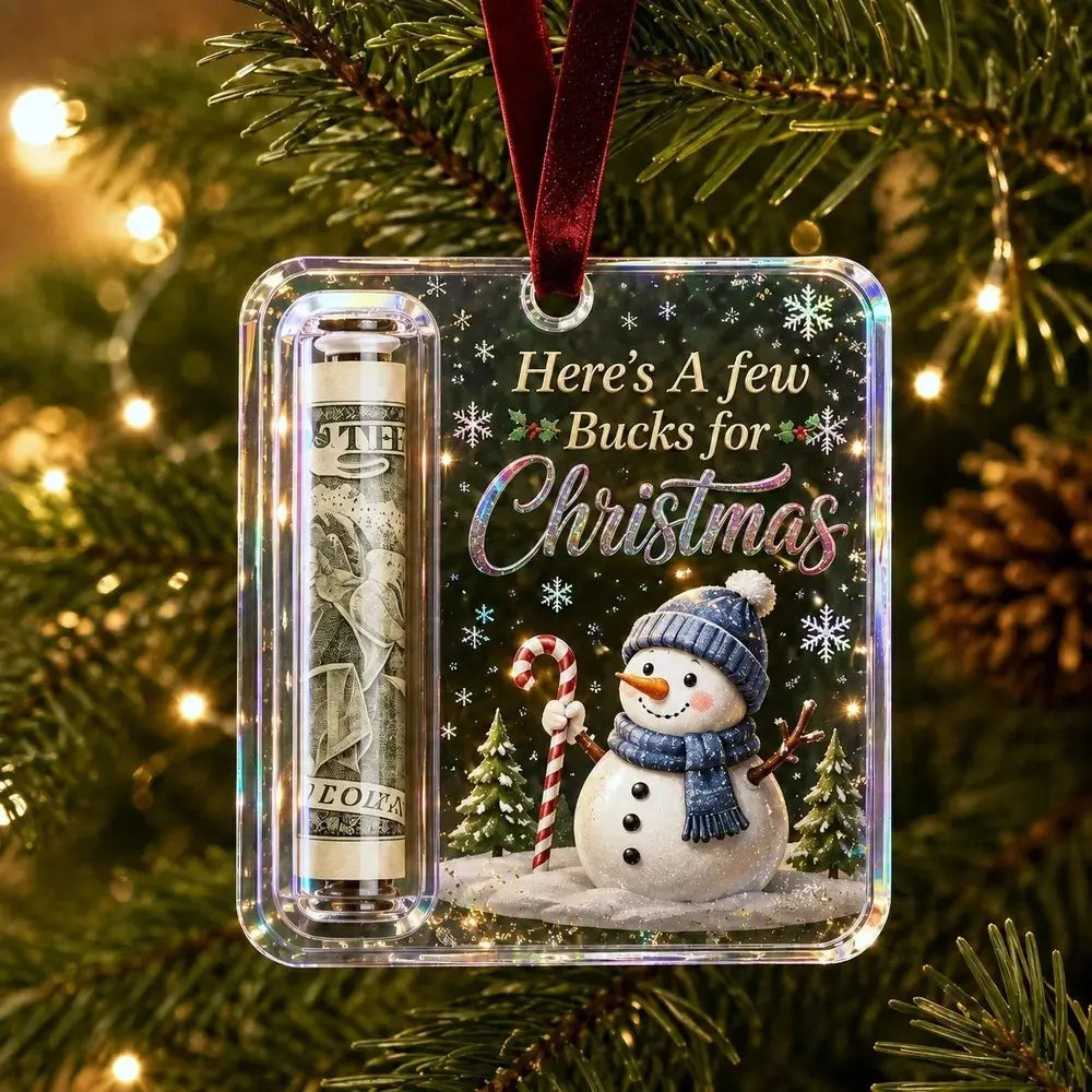 A personalized Christmas ornament hanging on a decorated Christmas tree. The clear square ornament contains a $100 bill inside a roll-up money tube and features a printed holiday design with a snowman, candy cane, pine trees, and the text 'Here's A few Bucks for Christmas'.