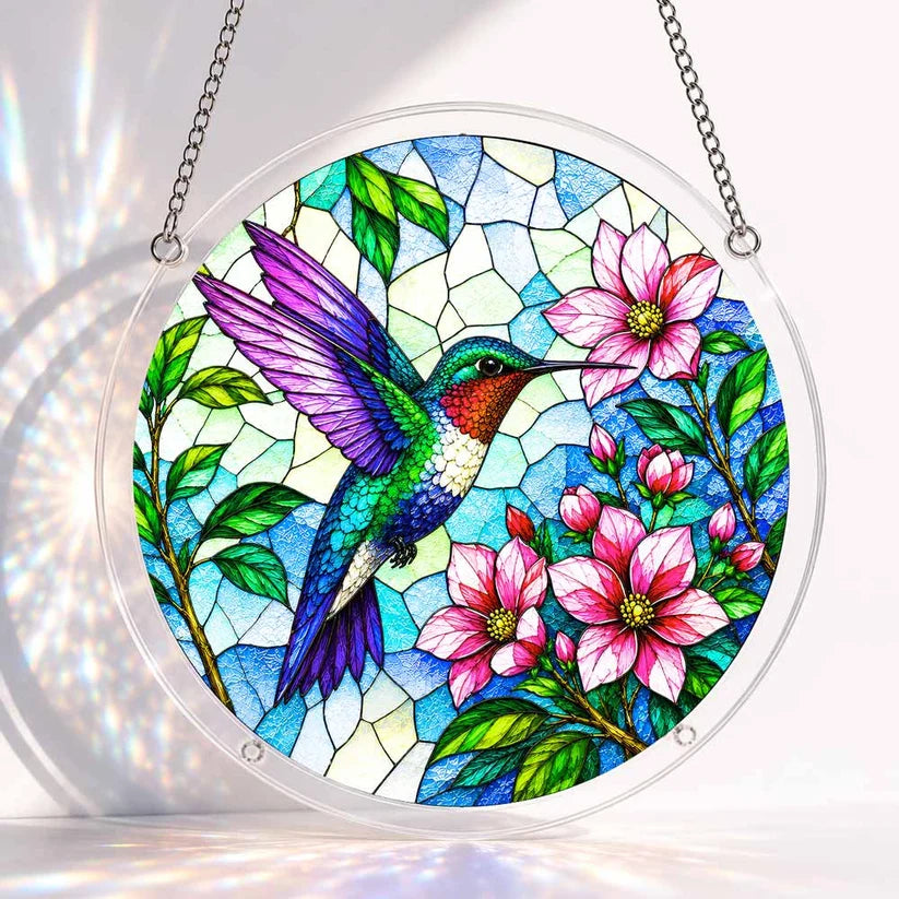 A round acrylic suncatcher featuring a colorful hummingbird beside pink blossoms on a blue stained glass background