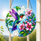 A round acrylic suncatcher with a colorful hummingbird beside pink blossoms hanging from a chain in front of a bright white window