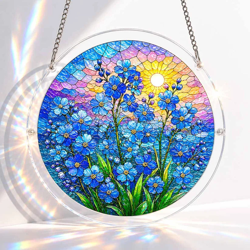 A round acrylic suncatcher with blue forget me not flowers and a glowing sun over a sunset sky in stained glass style