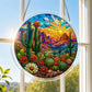A round acrylic suncatcher with saguaro cacti blooming flowers and desert mountains hanging from a chain in front of a bright white window