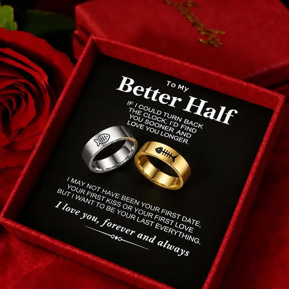 A gift box containing two rings, one silver and one gold, displayed on a black message card with romantic text. Red roses and a red fabric background surround the box.