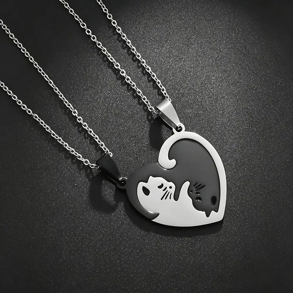 Two silver heart-shaped pendant necklaces on matching chains, designed to fit together into one heart. One pendant is polished silver with a black cat silhouette, and the other is black with a silver cat silhouette, set against a dark textured background.