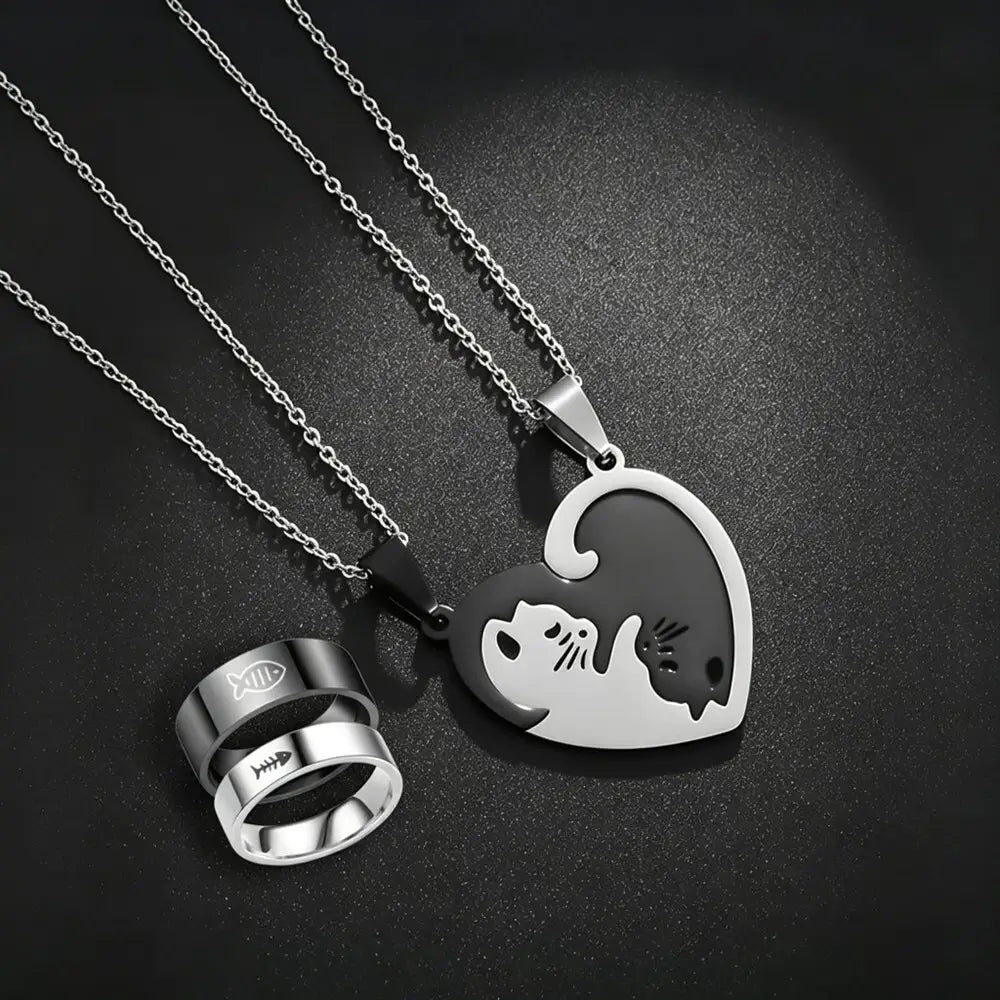Two silver-toned necklaces and matching rings displayed on a dark textured background. One necklace has a heart-shaped pendant split into white and black halves forming a cat and dog silhouette.