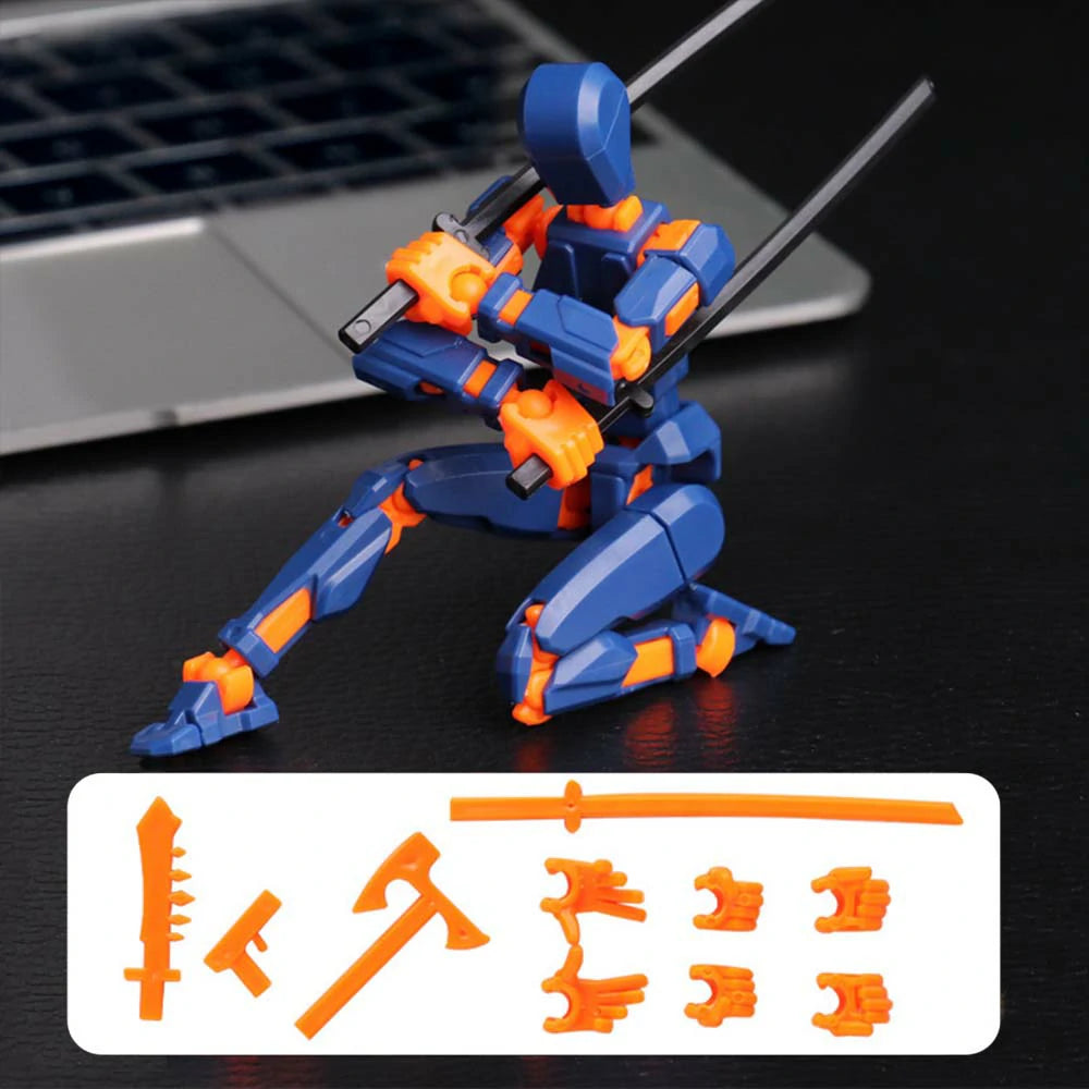 A blue and orange plastic robot action figure posed kneeling on a dark tabletop with a laptop blurred in the background. A white label at the bottom shows additional orange accessory pieces.