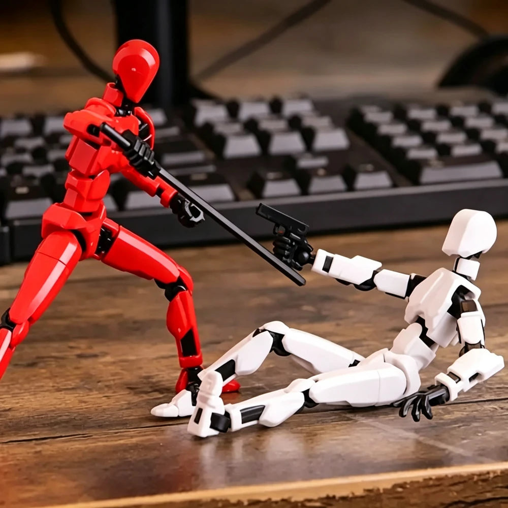 Two small humanoid action figures posed on a wooden tabletop with a blurred keyboard in the background. One figure is red and standing, holding a black gun.