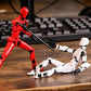 Two small humanoid action figures posed on a wooden tabletop with a blurred keyboard in the background. One figure is red and standing, holding a black gun.