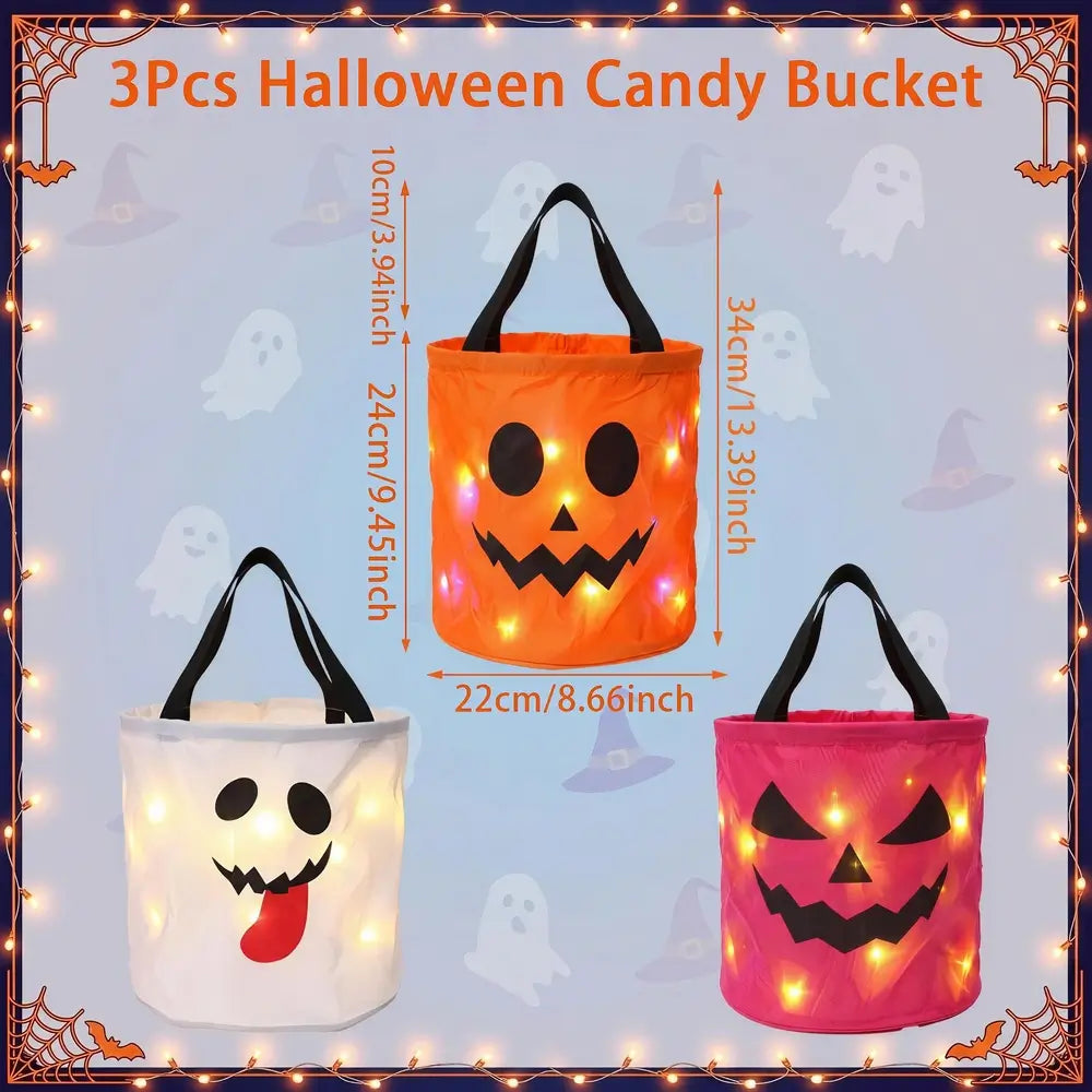 Three illuminated Halloween candy buckets shown on a light blue background with ghost and witch motifs, plus measurement labels. The top bucket is orange with a jack-o'-lantern face and black handles; the bottom left bucket is white with a ghost face and red tongue; the bottom right bucket is pink with a pumpkin face.