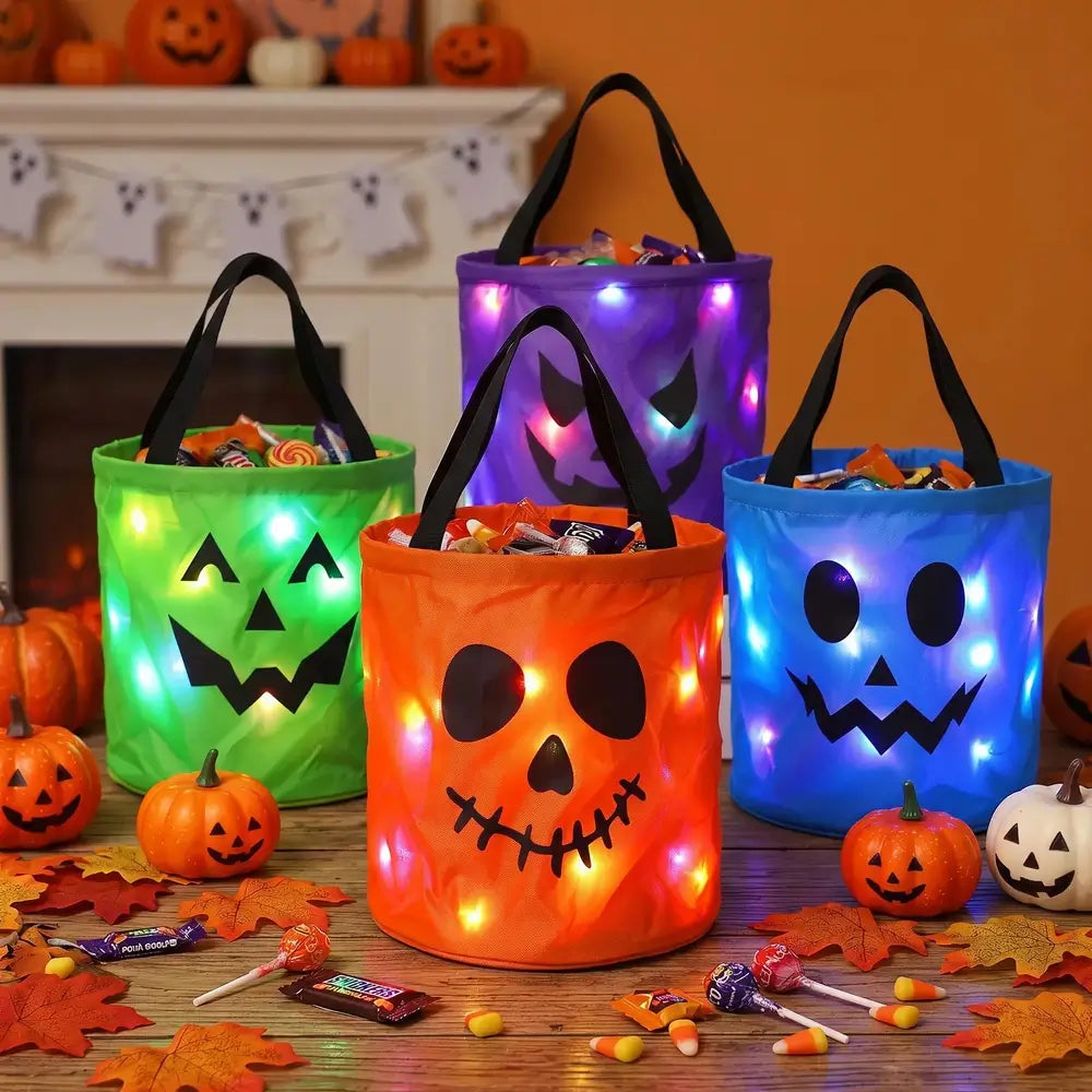 Four glowing fabric Halloween bucket bags in green, orange, purple, and blue, each printed with a jack-o'-lantern face and filled with candy, arranged on a table with small pumpkins and fall leaves in front of a fireplace.