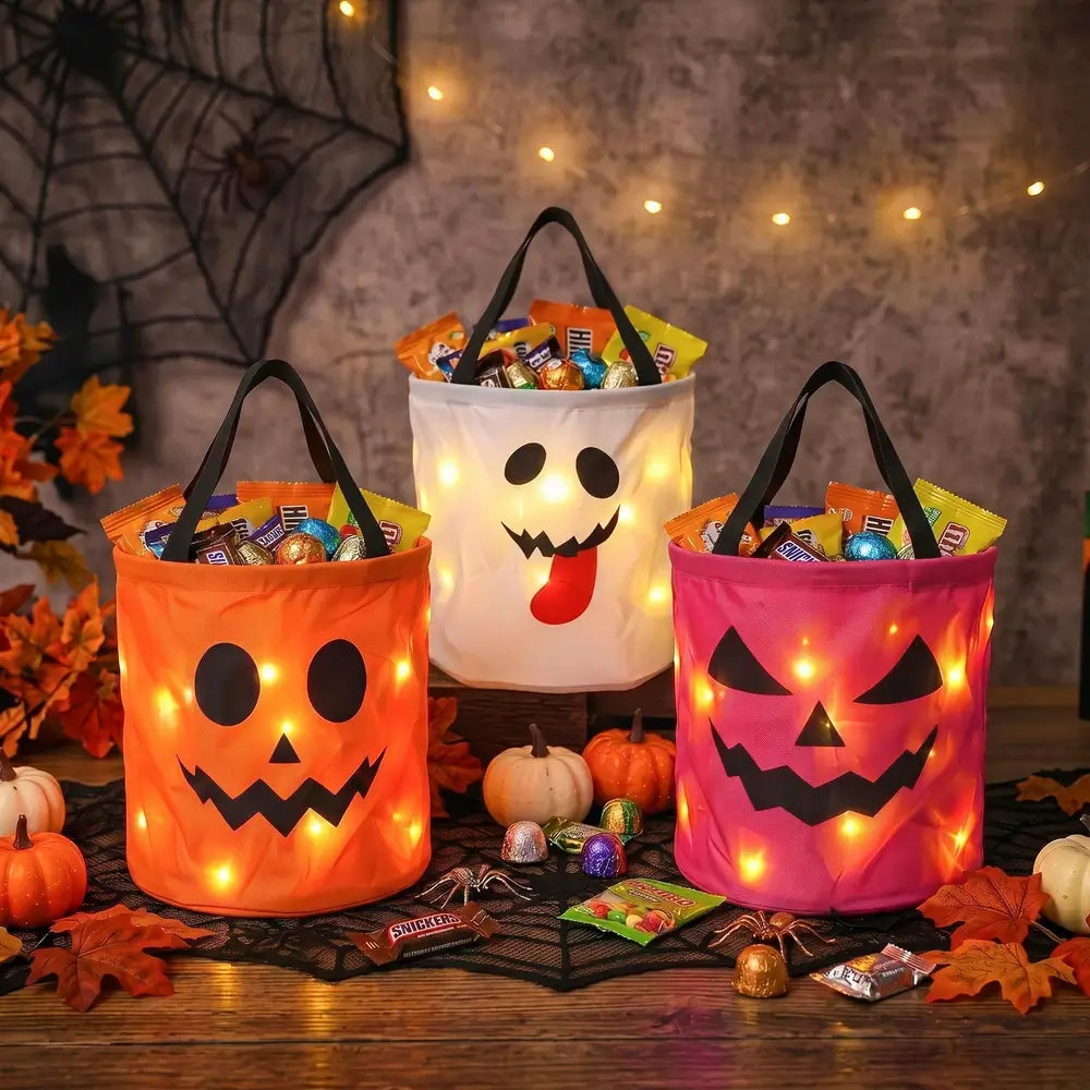 Three Halloween candy buckets with built-in string lights, decorated with jack-o'-lantern, ghost, and smiling faces, filled with assorted candy on a wooden table with autumn decorations.