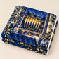 A folded Hanukkah blanket displays its golden menorah and olive branch watercolor design on a flat surface