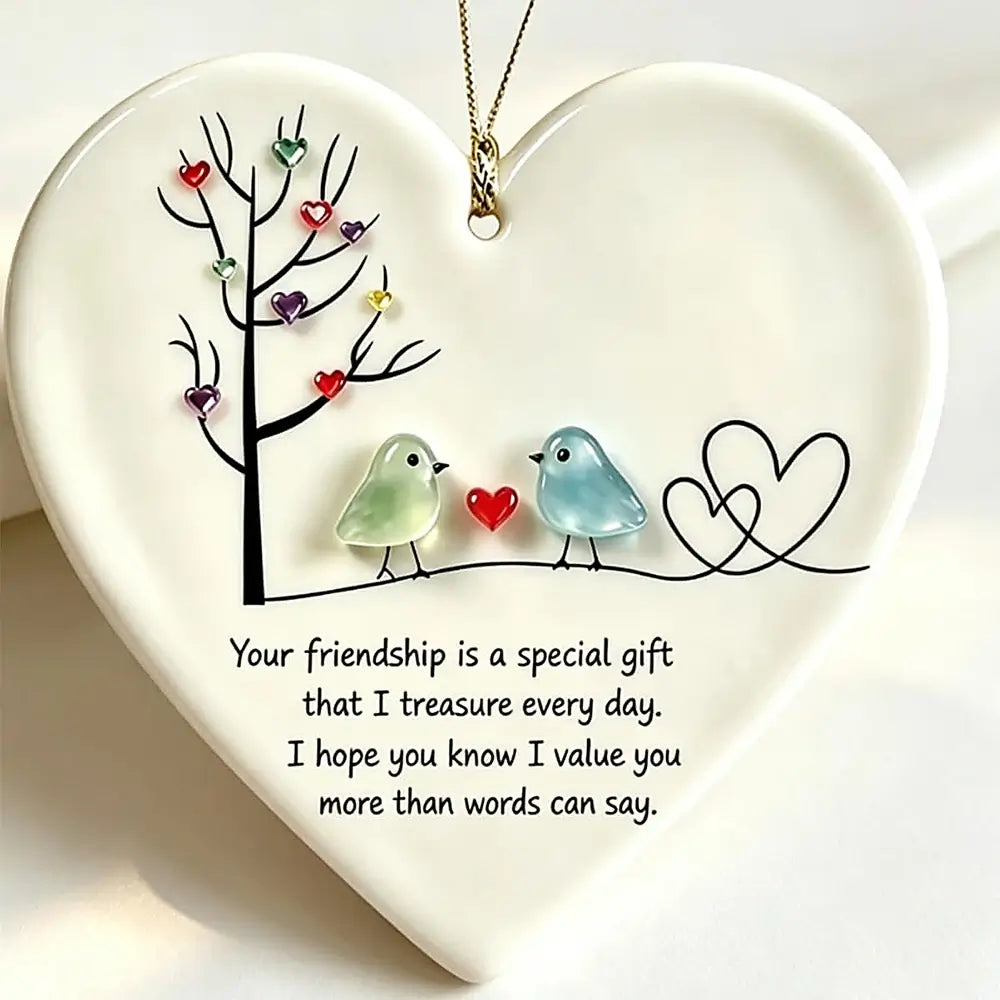 A heart-shaped ceramic ornament with a hanging loop, featuring a black tree with colorful heart-shaped leaves, two small birds facing each other with a red heart between them, and a black outlined double heart. Below the illustration is a printed friendship message.