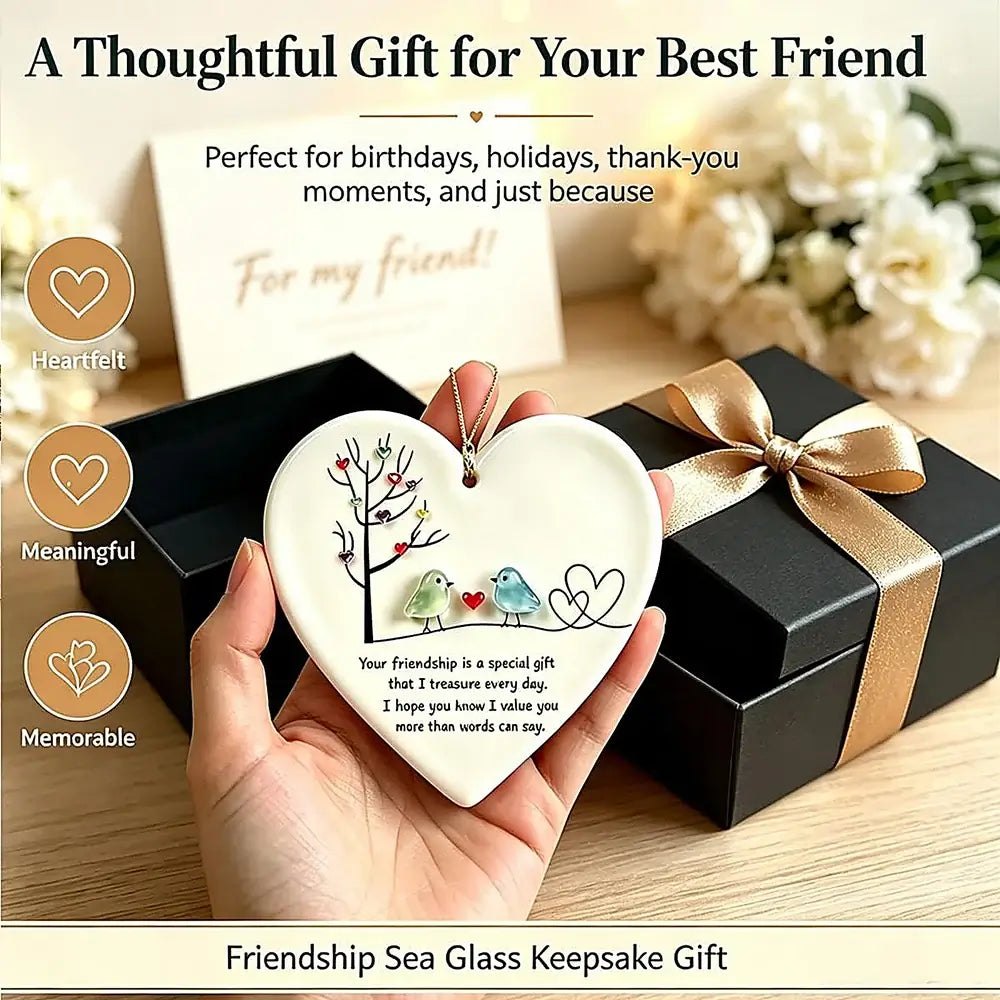 A hand holds a heart-shaped sea glass keepsake ornament featuring two birds and a tree, with a black gift box and ribbon in the background. Text overlays describe the thoughtful friendship gift.