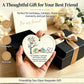 A hand holds a heart-shaped sea glass keepsake ornament featuring two birds and a tree, with a black gift box and ribbon in the background. Text overlays describe the thoughtful friendship gift.