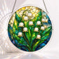 A round acrylic suncatcher showing white lily of the valley bells with green leaves and water droplets on teal glass