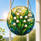 A round acrylic suncatcher with white lily of the valley bells and green leaves hanging from a chain in front of a bright white window