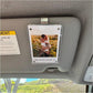A clear acrylic car sun visor photo holder mounted on a gray visor inside a vehicle. The holder contains a printed photo of a woman carrying a child in a field, with the text 'My entire world <3' at the bottom.