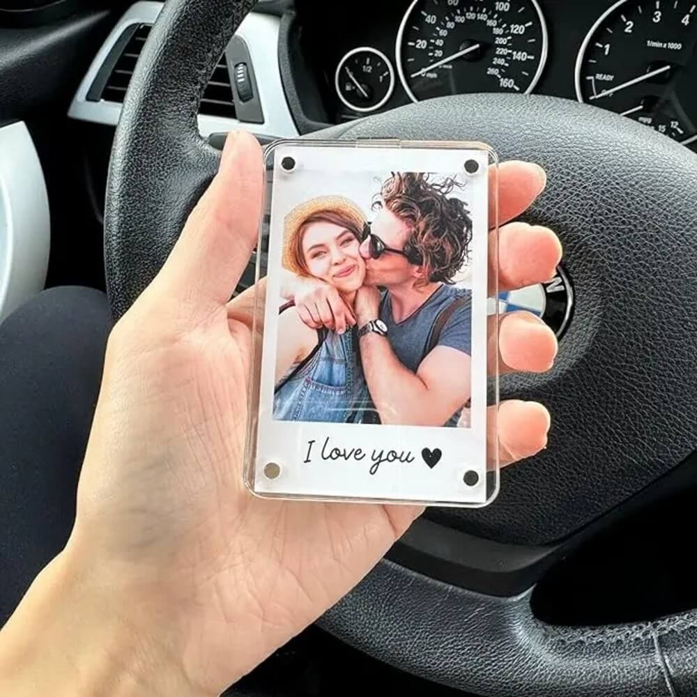 A hand holds a clear acrylic Polaroid-style photo frame in front of a car dashboard and steering wheel. The printed photo shows a couple hugging, with the text 'I love you' and a heart at the bottom.