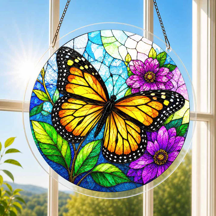 A round acrylic suncatcher with an orange black monarch butterfly and purple flowers hanging from a chain in front of a bright white window
