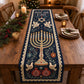 A navy quilted Hanukkah table runner printed with menorah, dove and pomegranate patterns laid on a wooden table decorated with pine branches and glowing candles