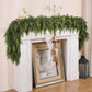 A green artificial pine garland draped over a white fireplace mantel, with a white knit stocking and gold bell hanging from the center. A framed winter landscape print leans in the fireplace, and a black lantern sits on the wooden floor against a pale pink wall.