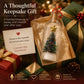 A holiday-themed gift presentation featuring a glass keepsake bottle with a miniature decorated Christmas tree and gold star, shown inside an open green gift box. Surrounding it are wrapped presents, pine branches, and warm holiday lights, with promotional text reading 'A Thoughtful Keepsake Gift' and feature icons at the bottom.