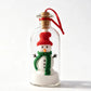 A clear glass bottle ornament featuring a small knitted snowman wearing a red hat and green scarf, sitting in white faux snow with a cork stopper and red hanging ribbon.