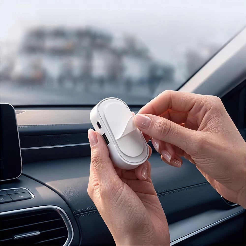A person's hands holding a small white oval car accessory with a peeling cover in front of a car dashboard and windshield.