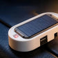 A close-up of a white portable solar power bank with a built-in solar panel on top, featuring a red power indicator light and USB-C port, resting on a dark textured surface.