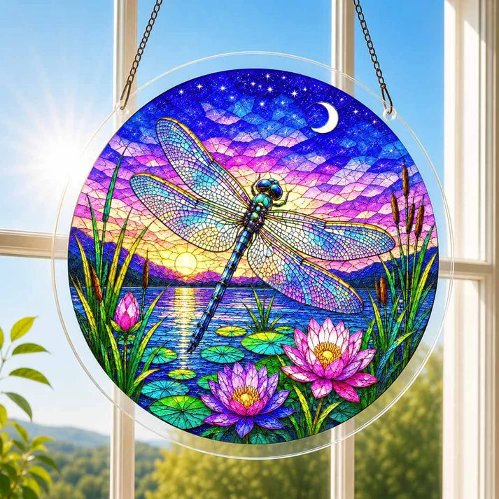A round acrylic suncatcher with a dragonfly over lotus leaves and flowers hanging from a chain in front of a bright white window