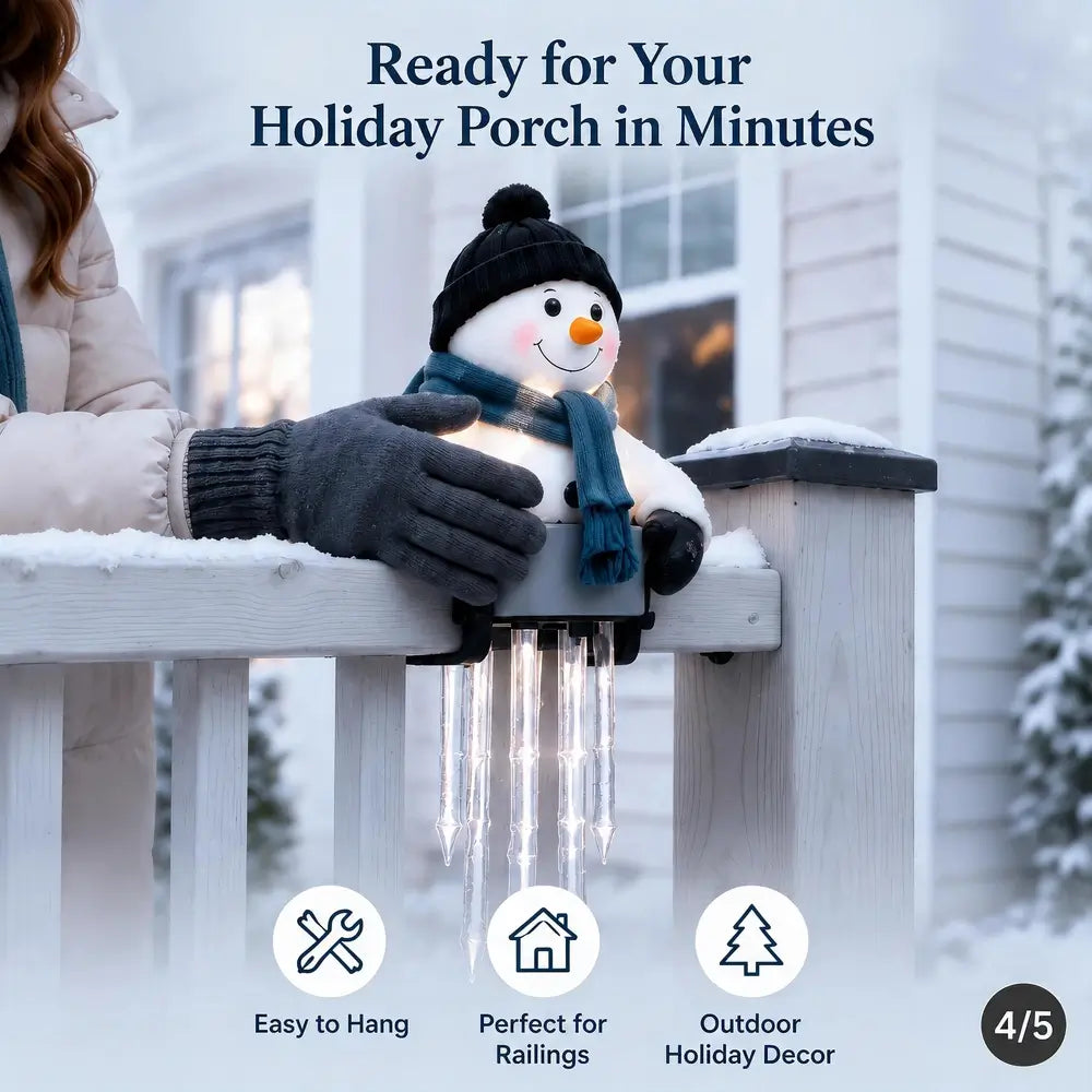 A snowman-shaped holiday porch light with a black hat, blue scarf, and glowing face is clipped onto a white railing in a snowy outdoor setting. A person's gloved hand is holding it in place.