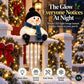 A plush snowman holiday light decoration sits on a snowy white porch railing decorated with pine garlands and warm string lights. The snowman wears a black hat and blue scarf and has glowing icicle accents hanging below.