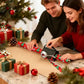 A man and a woman are wrapping a gift box with festive red, green, and white holiday wrapping paper on a granite countertop. The scene is decorated with a Christmas tree, wrapped presents, pinecones, and ornaments, creating a warm holiday atmosphere.